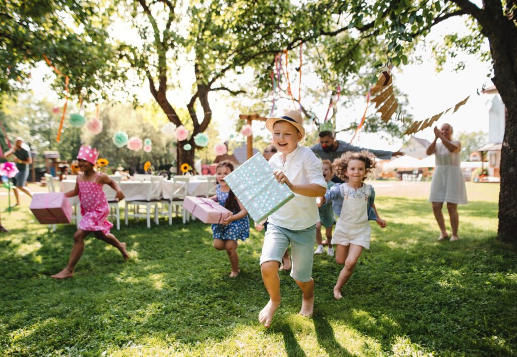 Kinder rennen lachend mit Geschenken durch den Garten bei Geburtstagsfeier mit einem Gartenpavillon